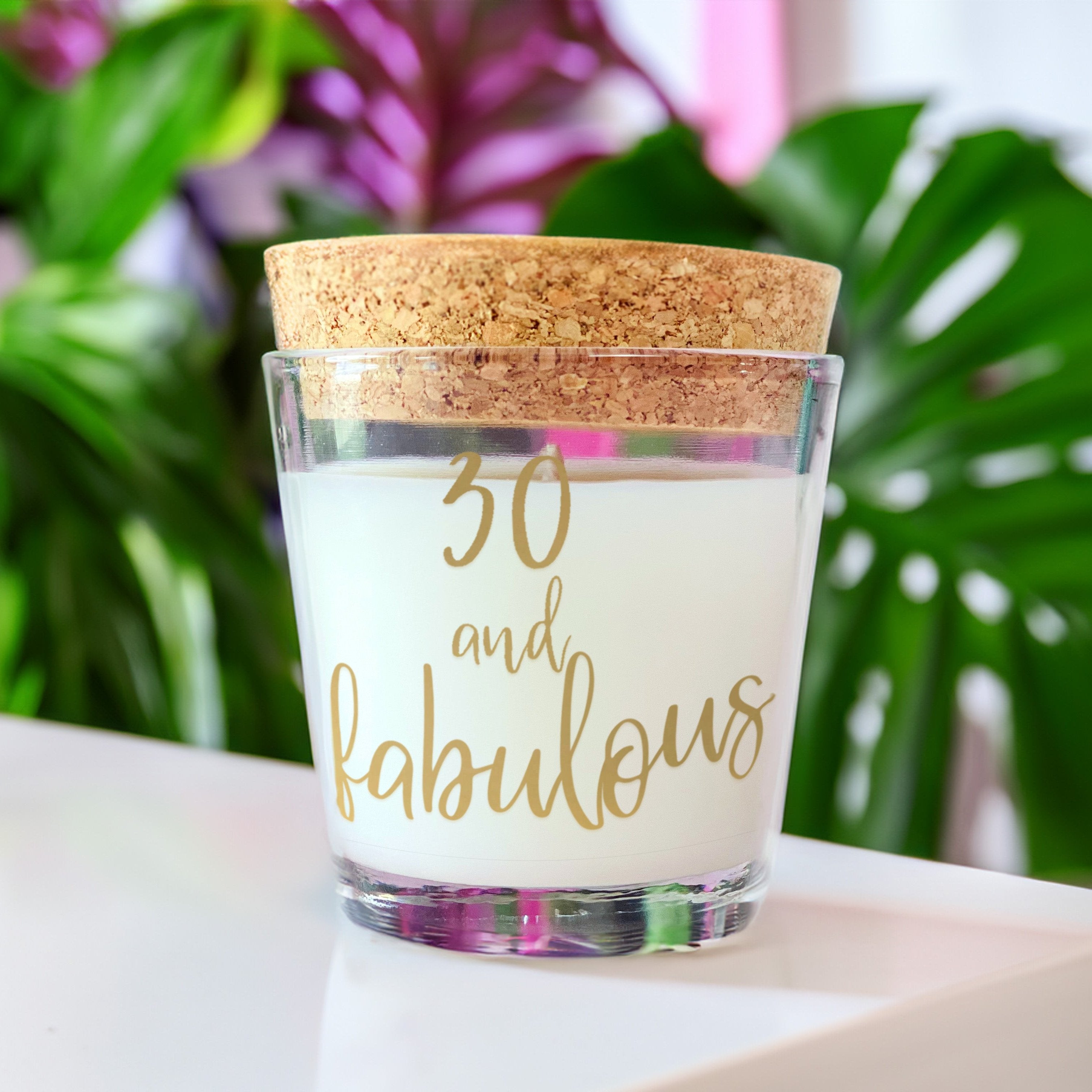 30 and fabulous candle with gold writing. Candle is white with cork lid. On a white table with green and purple blurred tropical leaves in background.