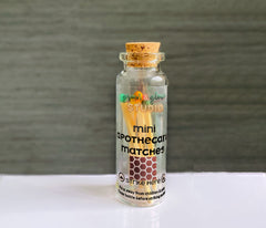 Mini Apothecary match bottle with cork lid. It has a clear label and match striker on front. On a white table with black linen background.